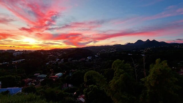 Ciel de feu rose et orange au-dessus de la ville. Le soleil couchant cr&eacute;e un contraste saisissant avec la silhouette urbaine et la v&eacute;g&eacute;tation luxuriante. Cr&eacute;puscule d'une intensit&eacute; sublime.