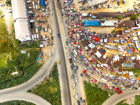 Aerial view of a bustling market meeting the highway, cars stream by amidst vibrant stalls and buildings casting shadows, Kilometer 16, Port Harcourt, Rivers State, Nigeria.