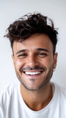 Fototapeta premium Happy young man with dark curly hair and beard smiles showing teeth looking directly at camera.