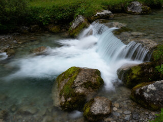 Fototapeta premium a small waterfall in the mountains of the alps, water flowing over moss-covered rocks into a crystal-clear mountain stream with long exposure