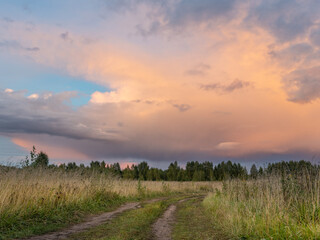 Road in a field with a cloudy sky and a rainbow in the distance