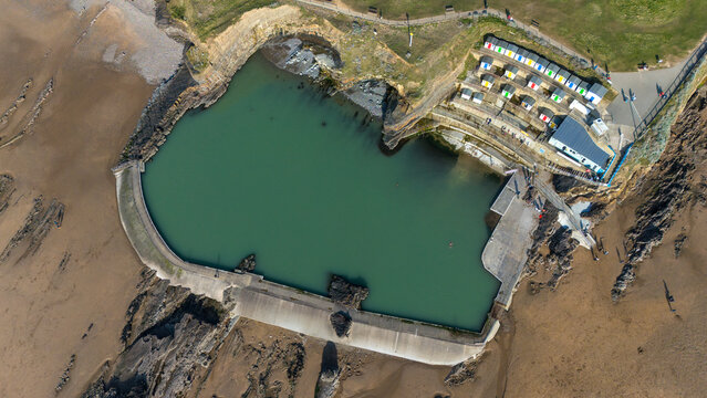 Aerial view of the vibrant turquoise water of Bude Sea Pool enclosed by dark rocky cliffs and sandy shores, with colorful beach huts adding a playful contrast, Bude, England, United Kingdom.