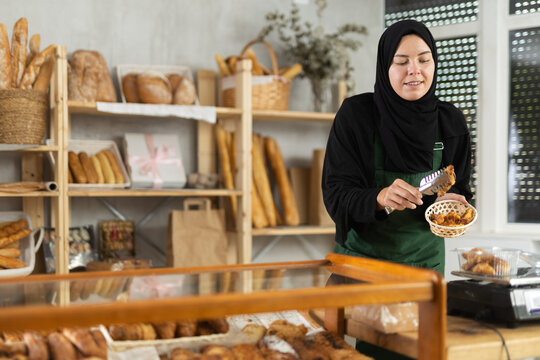 Muslim girl is working in sales area of bakery near counter, selling pastries. She puts croissants in disposable bag, packs goods, prepares order for buyer