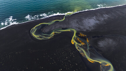 Aerial view of a serpentine river snaking through black volcanic sand, its waters swirling with ochre and jade hues as it meets the turquoise sea, Hvolsvollur, Rangarbing eystra, Iceland.