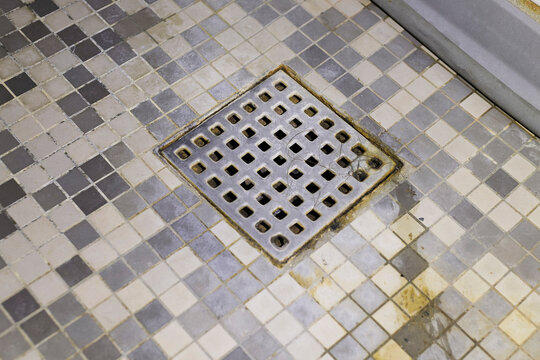 A close-up view of a metal square shower drain and grimy checkerboard mosaic floor tiles showing discoloration, mold, and trapped hair.