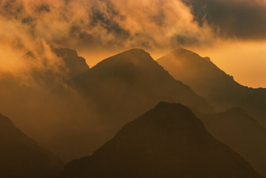 Aerial view of the rugged peaks of the Western Tatras pierce through a swirling veil of mist, their silhouettes painted in warm hues by the setting sun, Jamnik, Zilina Region, Slovakia.