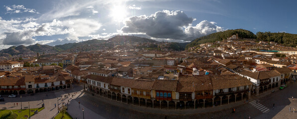 Panoramic view of colonial rooftops and arcades in Cusco, Peru, featuring a bright sunburst,...