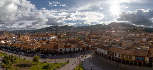 Obraz premium Scenic panorama of the Plaza de Armas in Cusco, Peru, featuring colonial arcades, red tile roofs, and hillside neighborhoods under a dramatic sunny sky with clouds.