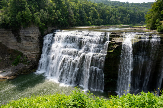 Lush green landscape surrounding a beautiful waterfall cascading down rocky cliffs into a serene lake on a clear sunny day in nature's embrace - Powered by Adobe