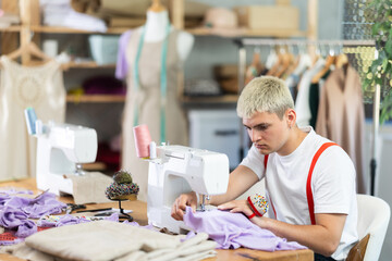 Attentive man tailor sewing textile using sewing machine sitting at workplace in small fashionable...