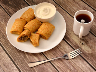 Pancakes stuffed with minced meat and sour cream, a cup of hot black coffee and a fork on a wooden table.