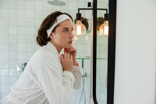 Woman wearing bathrobe and headband in bathroom leaning and examining face in black-framed mirror