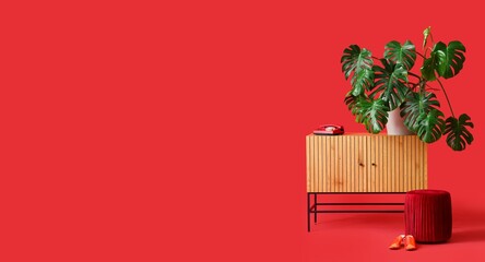 Commode with Monstera plant, telephone, pouf and female shoes on red background with space for text