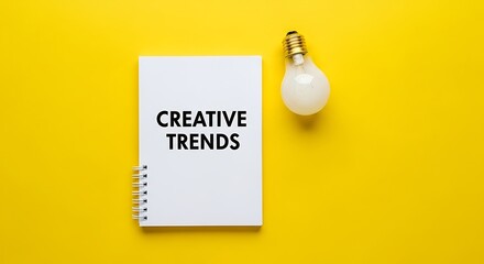 Top view of a white notebook with creative trends written on it next to a light bulb on yellow background