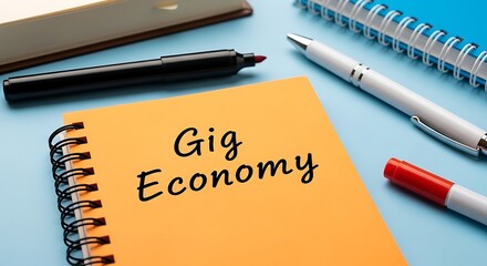 Orange notebook with gig economy handwritten on it surrounded by various pens and office supplies on a blue surface