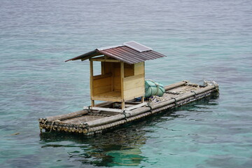 A simple, homemade bamboo raft with a small wooden shelter and a solar panel floating on clear, turquoise ocean water.