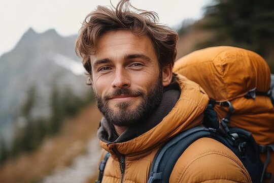 Smiling young man with beard wearing orange jacket and backpack standing outdoors with mountain background