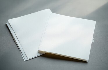 A stack of plain white blank textured paper sheets placed on a smooth gray surface with soft natural light casting gentle shadows