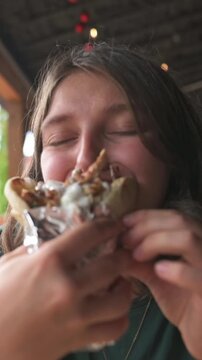 young woman biting wrapped sandwich at rustic outdoor eatery, closeup of hands and saucy filling with warm string lights overhead, eyes closed in bliss, candid foodie scene with fries and lively patio