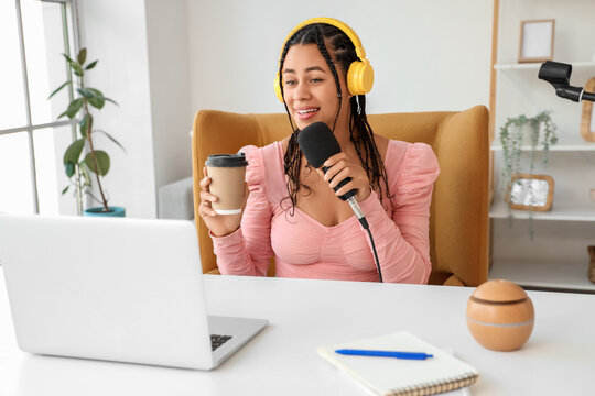 Young African-American woman with coffee cup and microphone recording podcast in studio