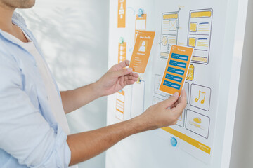 A designer is developing UX UI layouts, reviewing mobile app prototypes, and arranging interface elements on paper and board. He is focused on user experience and digital creativity.