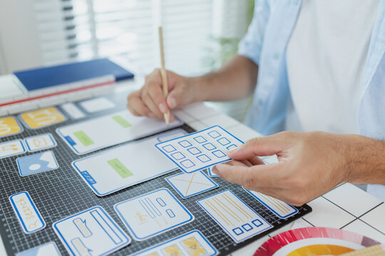 A designer is developing UX UI layouts, reviewing mobile app prototypes, and arranging interface elements on paper and board. He is focused on user experience and digital creativity. - Powered by Adobe