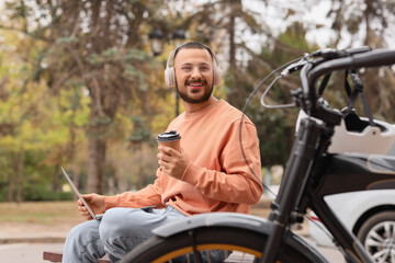 Young happy man in headphones with laptop, cup of coffee and bicycle sitting on bench outdoors