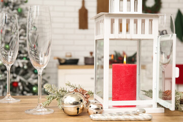 Lantern with burning candle, Christmas bells and glasses on table in kitchen