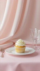 Vanilla cupcake with white buttercream frosting on a white plate, set on a pink tablecloth with draped pink fabric in the background and two empty wine glasses.
