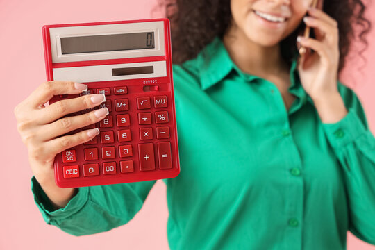 Young African-American woman with calculator talking by mobile phone on pink background - Powered by Adobe