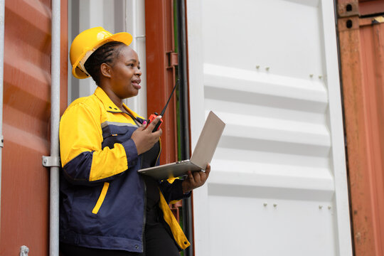 Female industrial worker communicating via walkie talkie while using laptop at container yard, Confident dock worker managing logistics with laptop and radio at shipping terminal - Powered by Adobe