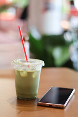 Refreshing iced matcha latte in plastic cup with red straw on wooden table next to smartphone. relaxing cafe scene with blurred background for moment of calm