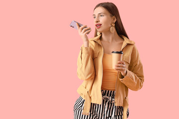 Beautiful young woman with cup of coffee and modern mobile phone recording voice message on pink background