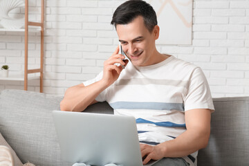 Handsome young man with laptop talking by mobile phone on sofa at home