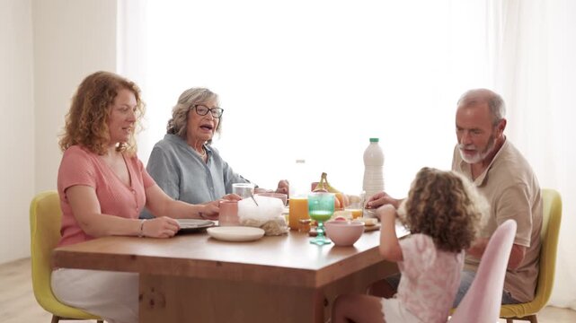 Happy three-generation family sitting at the dining table, eating and talking. Grandparents sharing a meal with their daughter and granddaughter