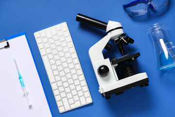 Modern microscope with keyboard, flask, clipboard and safety glasses on blue background