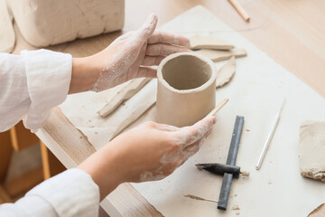Young female potter shaping clay pot with wooden rib at table in home studio, closeup