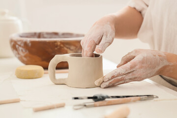 Young female potter shaping clay cup with hands on table in home studio, closeup