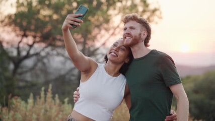 Young mixed-race couple happily taking a selfie with a smartphone while hugging in nature. Millennial man and woman creating content outdoors