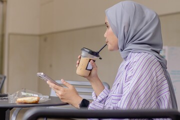 A young Asian Muslim woman enjoying her iced coffee while waiting for a call from someone