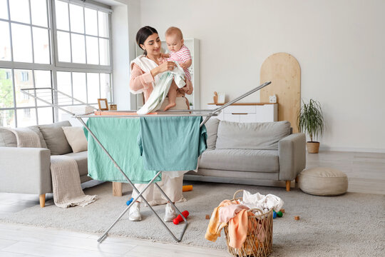 Young mother with cute little baby hanging laundry on drying rack at home