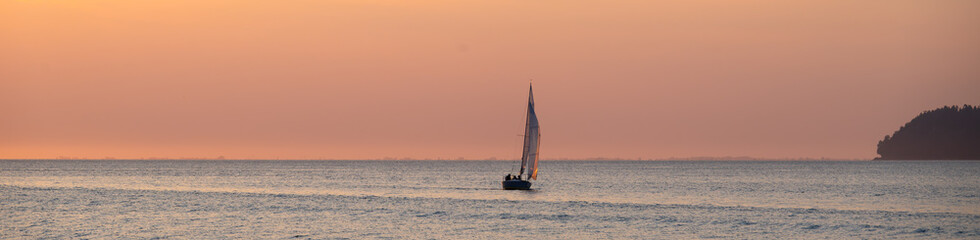 Fototapeta premium Peaceful recreation sailing in of the Pacific Coast in small sailboat at sunset, pastel sky in yellow an orange against blue water 