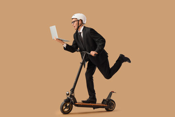 Young businessman with laptop riding electric scooter against beige background