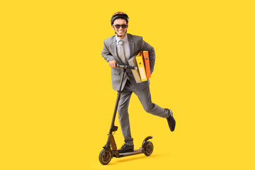 Young businessman with office folders riding electric scooter on yellow background