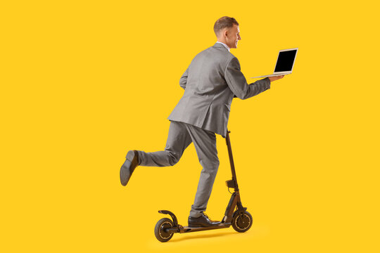 Young businessman with laptop riding electric scooter on yellow background