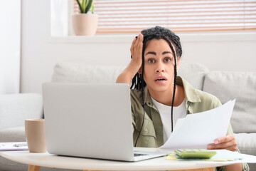 Shocked young African-American woman with tax form and laptop paying taxes online at home