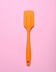 A bright orange silicone spatula placed upright on a soft pastel pink background, perfect for baking and cooking themes.