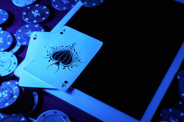 Tablet computer with poker chips and playing cards on blue table in casino, closeup