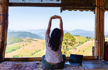 Digital nomad woman taking a wellness break, stretching with arms raised on a balcony overlooking mountain view with laptop nearby. Concept of work-life balance, mindfulness, and remote work.