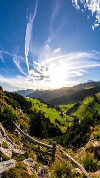 Sunny landscape photograph featuring a vibrant valley with rolling green hills beneath a bright blue sky filled with wispy clouds